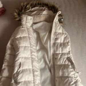 Vit dunjacka från H&M - Säljer en snygg vit dunjacka från H&M med pälskantad huva. Jackan har dragkedja och är quiltad för extra värme. Perfekt för kyliga dagar!