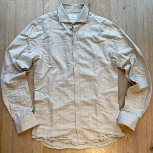 Beige skjorta från The Shirt Factory - Snygg beige skjorta från The Shirt Factory i slim fit. Skjortan har långa ärmar och knappar framtill. Perfekt för en stilren look. Materialet är lite linneliknande, men lite tjockare. Väldigt mjukt och skönt. Det står storlek 38-de motsvarar drygt en storlek M. Skriv vid frågor!