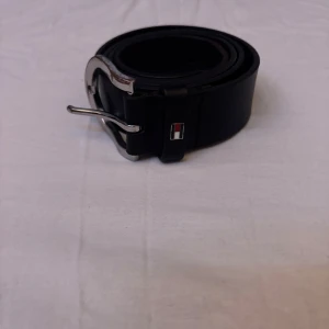 Svart Tommy Hilfiger bälte (Pris kan diskuteras!) - Snyggt svart bälte från Tommy Hilfiger med en stilren silverfärgad spänne. Bältet har en diskret logotypdetalj i rött, vitt och blått. Perfekt för att ge en elegant touch till din outfit. 