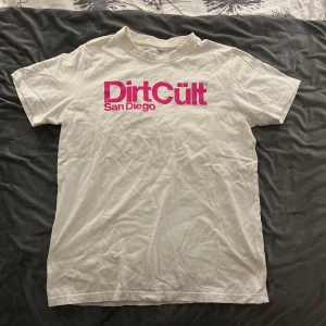Vit t-shirt från DirtCült - Säljer en vit t-shirt från DirtCült med rosa tryck 'San Diego' på framsidan. T-shirten är kortärmad och har en klassisk passform. Perfekt för en avslappnad stil.