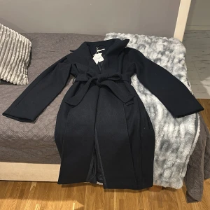 Marinblå kappa från Gina tricot - Elegant marinblå kappa från Gina tricot i storlek S. Kappan har ett stilrent bälte i midjan med långa ärmar och lång i längden. Perfekt för kyligare dagar med sin klassiska design.