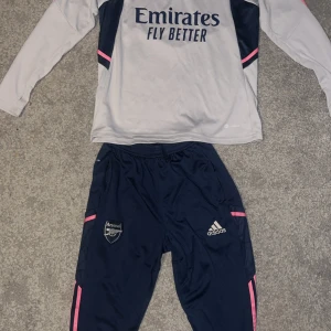 Träningsset från Adidas och Arsenal - Snyggt träningsset från Adidas med Arsenal-logga. Tröjan är långärmad och vit med mörkblå detaljer och rosa inslag. Byxorna är mörkblå med rosa detaljer och har både Adidas- och Arsenal-loggor. Perfekt för fotbollsfans!