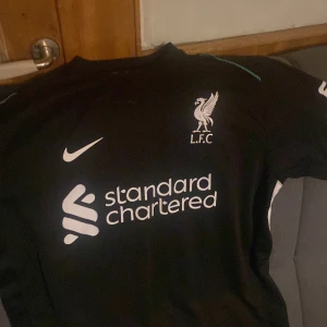 Liverpoll  - Snygg svart fotbollströja från Nike med L.F.C.-logga och Standard Chartered som sponsor. Tröjan har korta ärmar och en stilren design med vita detaljer. Perfekt för fotbollsfans som vill visa sitt stöd.