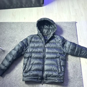 Svart dunjacka från Massimo Dutti - Snygg svart dunjacka från Massimo Dutti med huva och dragkedja. Perfekt för kyliga dagar med sin quiltade design som håller värmen. Jackan har en klassisk look och passar bra till olika outfits. han på bilden är 180 