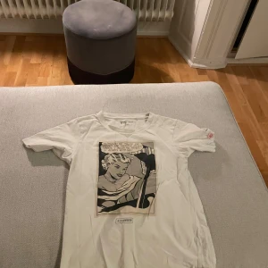 Vit t-shirt med tryck från Uniqlo - Säljer en vit t-shirt från Uniqlo med ett coolt tryck på framsidan i serietidningsstil. T-shirten har korta ärmar och en rund halsringning. Perfekt för en avslappnad stil.