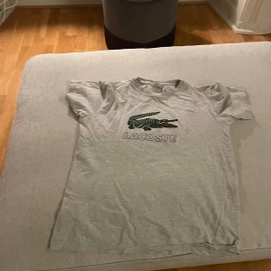Grå t-shirt från Lacoste - Säljer en grå t-shirt från Lacoste med det klassiska krokodilmärket på bröstet. T-shirten är kortärmad och har en normal passform. Perfekt för en avslappnad stil.