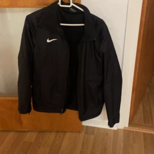 Svart jacka från Nike - Snygg svart jacka från Nike med dragkedja framtill och klassisk logga på bröstet. Perfekt för sportiga aktiviteter eller som en stilren vardagsjacka. Jackan har långa ärmar och en enkel design som passar till det mesta.