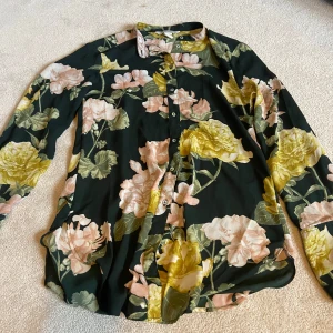 Blommig blus från H&M Conscious - Snygg blommig blus från H&M Conscious med stora gula och rosa blommor på en mörkgrön bakgrund. Blusen har långa ärmar och knappar framtill. Perfekt för att liva upp garderoben med lite färg och mönster💞