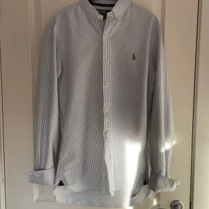 Randig skjorta från Polo Ralph Lauren - Snygg och stilren randig skjorta från Polo Ralph Lauren i slim fit. Skjortan är vit med blå ränder och har en broderad logotyp på bröstet. Perfekt för en elegant look. Skjortan är använd 5-7 gånger 😊(Skjortan passar mig som är 172 CM)