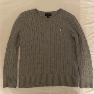 Grå kabelstickad tröja från Ralph Lauren - Säljer en stilren grå kabelstickad tröja från Ralph Lauren i stolek L(14-16). Tröjan har en klassisk design med rund hals och det ikoniska broderade logotypen på bröstet. Perfekt för kyligare dagar.