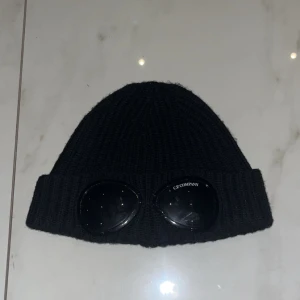 C.P Company mössa - Snygg svart ribbad mössa från C.P. Company med unika glasögondetaljer framtill. Perfekt för att ge din outfit en cool touch. Tillverkad i ett mjukt och bekvämt material, skick 9/10 (passar alla) hör av er vid minsta fråga.( köparen står för )frakt👊🏼😁