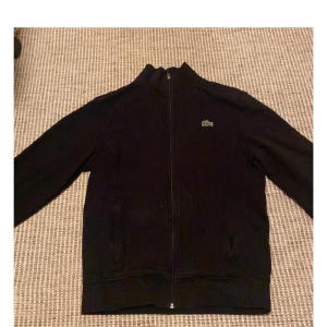 Svart jacka från Lacoste - Säljer en stilren svart jacka från Lacoste med dragkedja och det klassiska krokodilmärket på bröstet. Jackan har långa ärmar och en hög krage, perfekt för en sportig look.