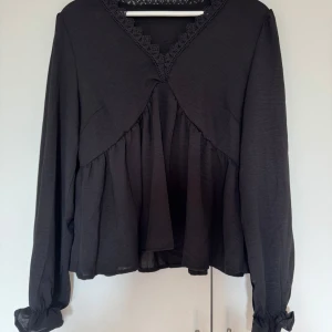 Svart blus med spetsdetaljer - Säljer en elegant svart blus med v-ringad spetsdetalj. Blusen har långa ärmar och en lös passform som ger en skön och avslappnad känsla. Perfekt för både vardag och fest!