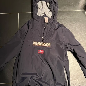 Mörkblå vindjacka från Napapijri - Säljer en snygg mörkblå vindjacka från Napapijri. Den har en praktisk huva och en halv dragkedja framtill. Jackan är perfekt för blåsiga dagar och har en cool logga med en norsk flagga på bröstet. Insidan är fodrad för extra komfort. Perfekt för höst och vår!