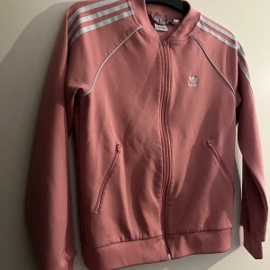 Adidas tröja - En rosa Adidas tröja med vita ränder och en liten logga på bröstet. Tröjan har en dragkedja framtill och två fickor på sidorna. Inga konstigheter, dragkedjans löpare har bara gått sönder men går att dra upp och ner som vanligt.