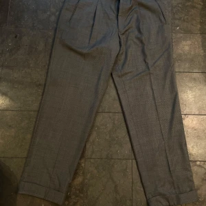Grå kostymbyxor - Säljer ett par stiliga grå kostymbyxor i klassisk design från Oscar Jacobsson. Byxorna har en rak passform med pressveck och uppvikta benslut. Säljer även tillhörande överdel av kavaj i annan annons.