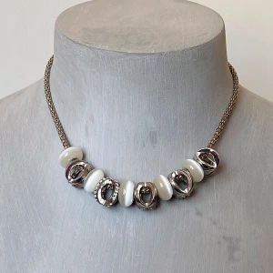 Elegant halsband med vita och silverdetaljer - Säljer ett elegant halsband med vita pärlor och silverfärgade detaljer. Kedjan är i en fin flätad stil och har en justerbar längd med ett hjärta som hänge baktill. Perfekt för att ge en stilren touch till din outfit!