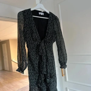 Svart blommig klänning med knyt - Säljer en superfin svart klänning med små vita gröna blommor. Den har en v-ringad omlott-design och knyt i midjan för en smickrande passform. Långärmad med ett lätt och luftigt material, perfekt för både vår och sommar. Klänningen är i nyskick och redo för nästa fest eller vardagsäventyr!