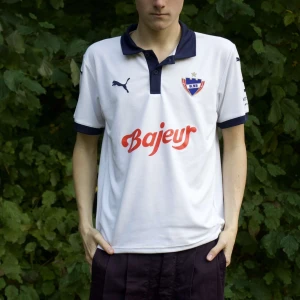  fotbollströja från b93 - Säljer en vit fotbollströja från Puma med mörkblå detaljer. Tröjan har korta ärmar och en krage i samma mörkblå färg. Den har tryck på bröstet och ärmen, samt en logga på ryggen. Perfekt för fotbollsfans eller som träningsplagg! B93 Danmark!! Nyskick