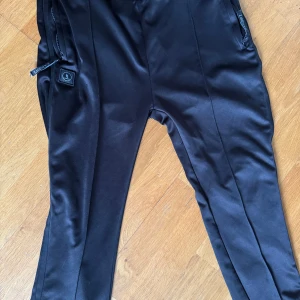 Svarta byxor från L'Agence - Snygga svarta byxor från L'Agence med dragkedjor vid fickorna. Byxorna har en elastisk midja för extra komfort och är tillverkade i ett glansigt material som ger en lyxig känsla.