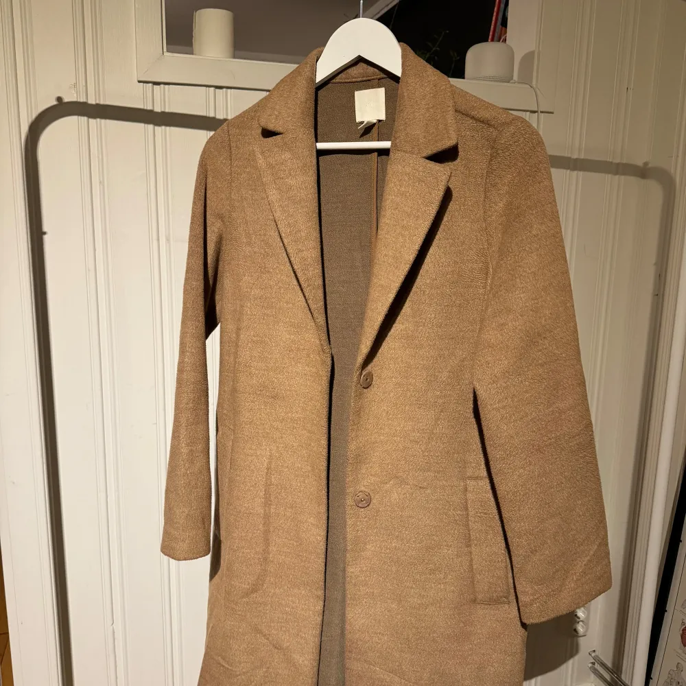 Snygg beige kappa från H&M i en klassisk design. Den har långa ärmar och knappar framtill. Perfekt för höst och vår, med en stilren look som passar till många tillfällen.. Takit.