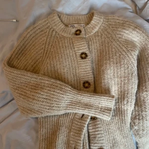 Beige stickad kofta från Zara - Mysig beige stickad kofta från Zara med stora knappar framtill. Perfekt för kyliga dagar med sin varma och bekväma design. Koftan har långa ärmar och en ribbad struktur som ger en stilren look.
