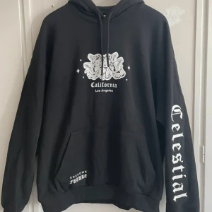 Svart hoodie från YOURTURN - Säljer en svart hoodie från YOURTURN med tryck av texten 'California Los Angeles' och 'Celestial' på ärmen. Den har en stor ficka framtill och justerbar huva med dragsko. Perfekt för en avslappnad stil.
