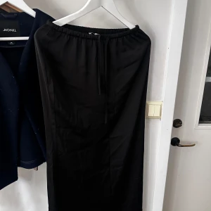 Svart satin långkjol från Nelly - Snygg svart satin långkjol från Nelly med elastisk midja för en bekväm passform. Perfekt för att skapa en stilren look. Kjolen är enkel och elegant, vilket gör den lätt att matcha med olika toppar.