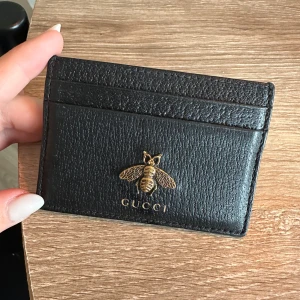 Svart kortfodral från Gucci - Säljer ett stilrent svart kortfodral från Gucci i skinn. Fodralet har en elegant guldfärgad bi-detalj och flera kortfack för praktisk förvaring. Perfekt för att hålla dina kort organiserade med stil.kan diskutera pris 