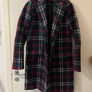 Rutig kappa - Meddellång rutig kappa från NA-KD i mörkblått, rött och vitt. Den har en klassisk krage och långa ärmar, perfekt för höst och vinter. Väldigt stilren design med knappar framtill. Något nopprigt då jag använt den väl men ser fortfarande bra ut