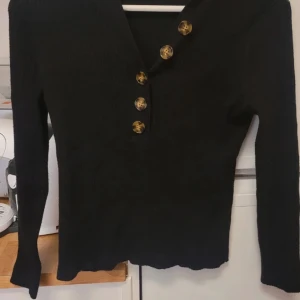 Svart tröja från KappAhl med knappar - Säljer en stilren svart tröja från KappAhl med dekorativa knappar framtill. Tröjan har en v-ringad design och långa ärmar, perfekt för en elegant look. Passar bra till både vardag och mer uppklädda tillfällen.