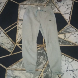 Grå mjukisbyxor från Nike - Snygga grå mjukisbyxor från Nike med elastisk midja och snörning. Byxorna har en liten Nike-logga på sidan och är perfekta för en avslappnad stil. De är tillverkade i ett mjukt material som ger hög komfort.