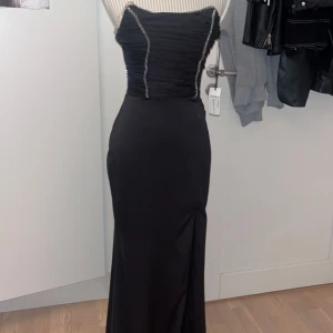 Svart axelbandslös långklänning - Elegant svart axelbandslös långklänning med glittriga detaljer längs bysten. Klänningen har en smickrande passform och en slits som ger extra rörelsefrihet. Perfekt för balen, köpt för 2500kr. Passar S/m aldrig använd har fortfarande prislappen