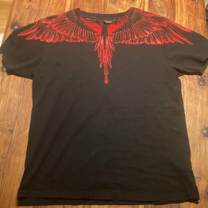 Svart t-shirt med rött vingmönster från Marcelo Burlon - Snygg svart t-shirt från Marcelo Burlon med ett iögonfallande rött vingmönster som sträcker sig över axlarna och ner på bröstet. Perfekt för dig som vill sticka ut med en unik design.