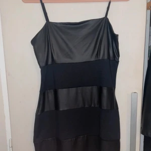 Svart axelbandslös klänning - Snygg svart klänning med axelband och blockmönster i olika material. Perfekt för en kväll ute eller en speciell tillställning. Klänningen har en stilren design med en mix av glansigt och matt tyg.