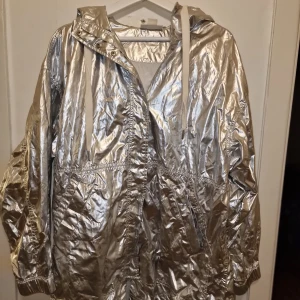 Silverfärgad jacka från Monki - Säljer en cool silverfärgad jacka från Monki. Jackan har en glansig finish och är utrustad med en huva och knappar framtill. Perfekt för att sticka ut i mängden!