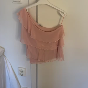 Rosa offshoulder topp med volanger - Säljer en söt rosa offshoulder topp med flera lager av volanger. Perfekt för en somrig look. Toppen har en asymmetrisk design som ger en unik stil.