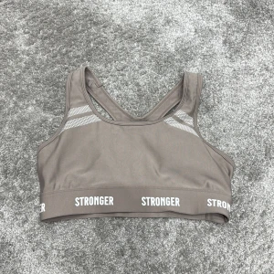 Beige sporttopp från Stronger - Snygg beige sporttopp från Stronger med bred resår och textdetaljer. Toppen har en racerback-design och är tillverkad i ett stretchigt material för optimal komfort under träning.