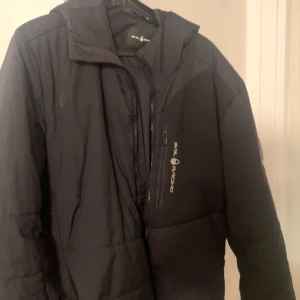 Svart dunjacka från Sail Racing - Säljer en svart dunjacka från Sail Racing med huva och dragkedja. Jackan har en logotyp på bröstet och en patch på ärmen med texten 'Sail Antarctica Expedition'. Perfekt för kalla dagar.