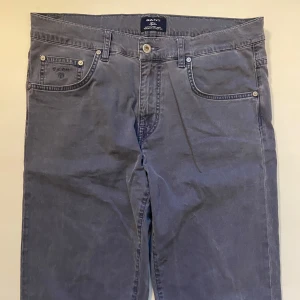 Grå byxor från Gant - Snygga grå byxor från Gant med klassisk femficksdesign och knappgylf. Byxorna har en stilren look med en läderpatch med logotyp på baksidan. Perfekta för en avslappnad men stilfull stil.