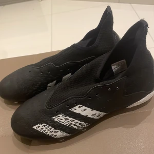 Svarta fotbollsskor från Adidas - Snygga svarta fotbollsskor från Adidas med vita detaljer och mönster. Skorna har en strömlinjeformad design med snörning och dobbar för bra grepp på planen. Perfekta för fotbollsträning och matcher.