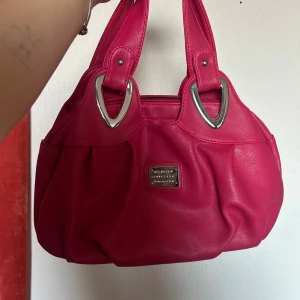 Rosa handväska med silverdetaljer - Snygg rosaröd handväska från Nine West i syntetmaterial. Väskan har stiliga silverdetaljer och en praktisk dragkedja upptill. Perfekt för att ge en färgklick till din outfit! Den är mellan stor!