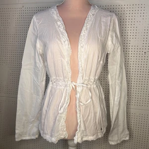 Vit kofta med spetsdetaljer - Säljer en söt vit kofta med spetsdetaljer och knytband i midjan. Koftan har långa ärmar och är perfekt för en romantisk look. Den är tillverkad i ett lätt och luftigt material.