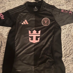 Svart fotbollströja från Adidas - Snygg svart fotbollströja från Adidas med rosa detaljer och klubbmärke på bröstet. Tröjan har korta ärmar och nummer 10 med namn på ryggen. Perfekt för fotbollsfans!