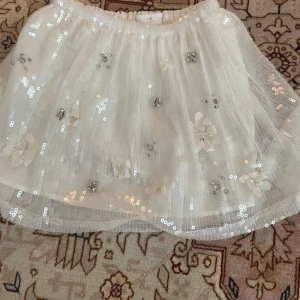 Vit paljettkjol från Zara - Säljer en superfin vit kjol från Zara med glittrande paljetter och blommiga detaljer. Kjolen har en elastisk midja och är perfekt för att lysa upp vilken outfit som helst. Den är kort och har ett lager av tyll som ger en extra volym. Perfekt för speciella tillfällen! ✨