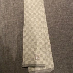 Grå rutig halsduk  - Snygg grå rutig halsduk i 100% ull. Den har ett diskret rutmönster och en elegant logotyp i ena hörnet. Perfekt för att hålla dig varm och stilren under kyliga dagar.