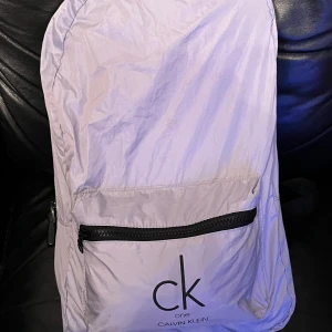 Vit ryggsäck från Calvin Klein - Säljer en stilren vit ryggsäck från Calvin Klein med svart dragkedja och logga. Perfekt för att bära dina grejer med stil. Ryggsäcken har justerbara axelremmar och en praktisk ytterficka med dragkedja.