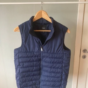 Mörkblå väst  - Säljer en snygg mörkblå quiltad väst från lyle and scott. Västen har en dragkedja framtill och är perfekt för lager-på-lager under kyligare dagar. Den har en stilren design som passar till många olika outfits.