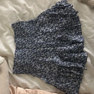 Blommig kortkjol i blått och vitt - Charmig kortkjol med blommigt mönster i blått och vitt. Kjolen har en volangdetalj. Är i storlek s