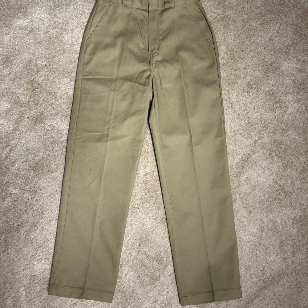 Snygga beige byxor från Dickies med en klassisk design. De har en rak passform och är tillverkade i ett slitstarkt material. Perfekta för en stilren look. Byxorna har dragkedja och knapp framtill.. Farkut & Housut.